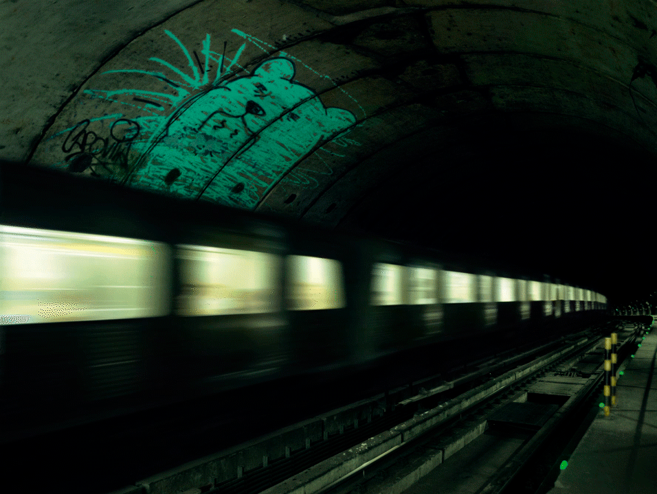 A train moving through a dark subway tunnel with green lighting. The walls of the tunnel are covered in graffiti
